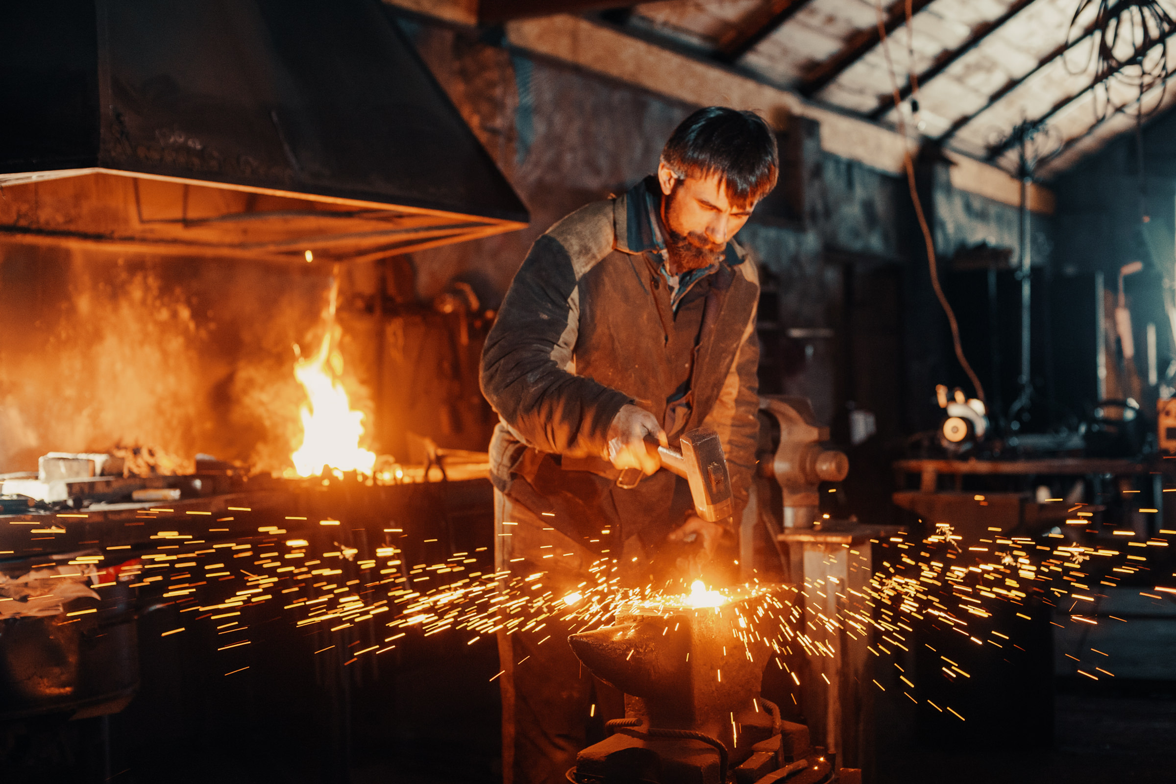 Фотография кузнеца за работой в кузнечном цеху возле наковальни с разлетающимися огненными искрами во время ковки металлической детали