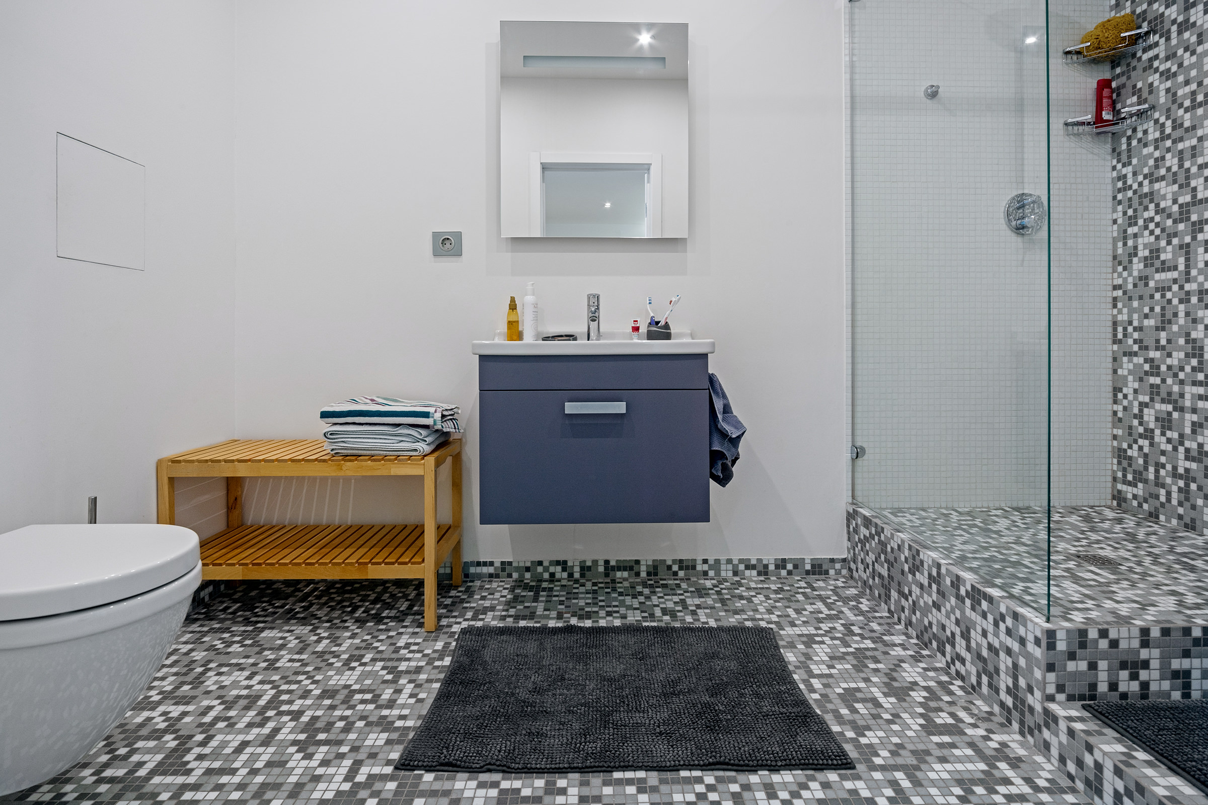 Интерьерная фотография совмещённой ванной комнаты с туалетом и душем в светлых тонах, мелкой серой плиткой и синей тумбочкой