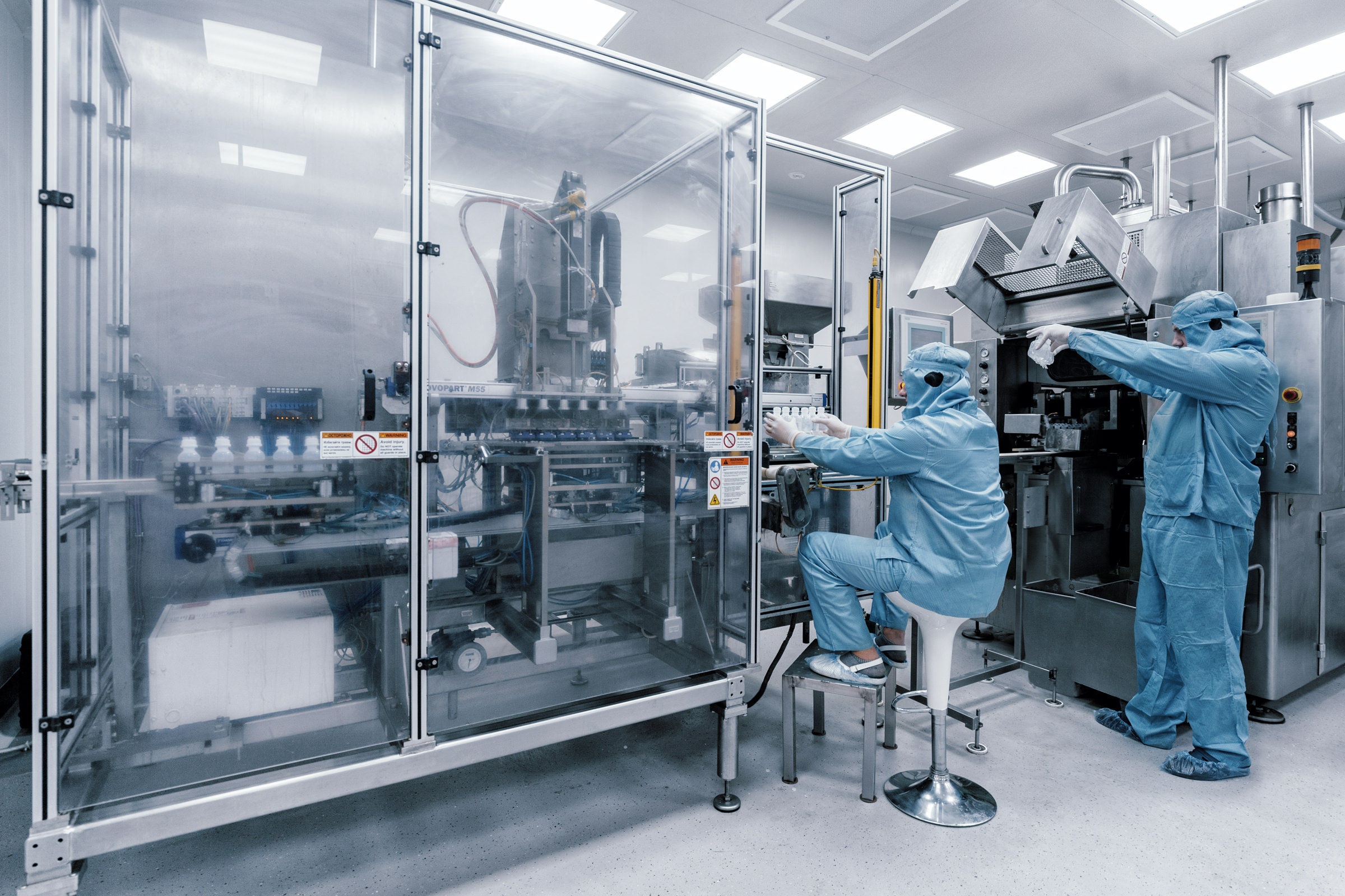 Индустриальная фотосъёмка линии по разливу медицинских препаратов с двумя лаборантами, контролирующими производственный процесс