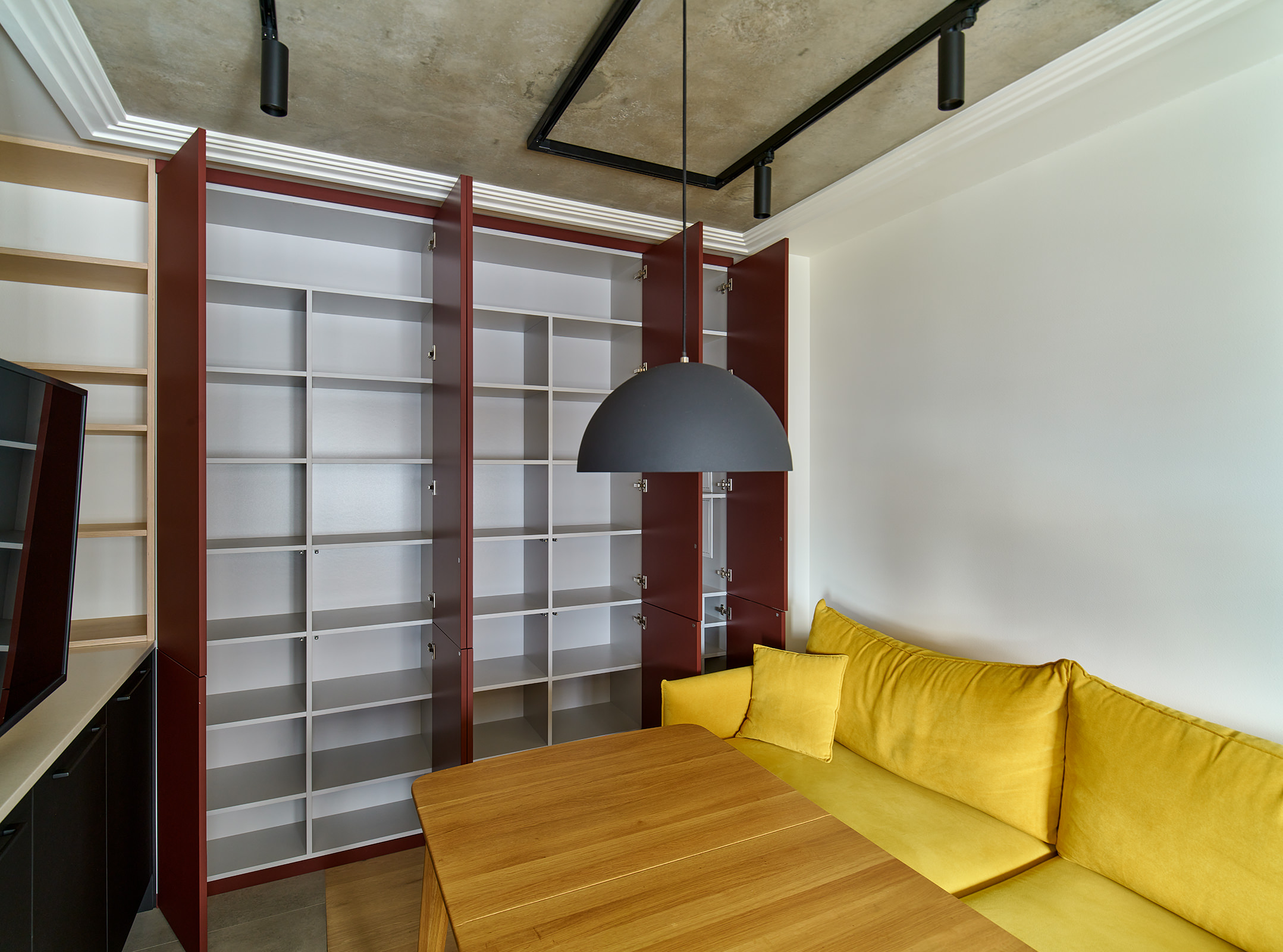 Фотография мебели и встроенного кухонного шкафа красно-бордового цвета с полками рядом с диваном горчично-желтого цвета и обеденным столом в интерьере новой квартиры в современном стиле